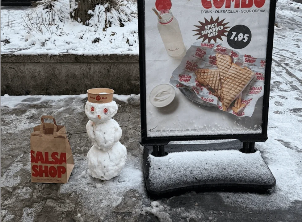 Salsa Shop team standing next to a snowman wearing a Salsa Shop to-go box as a hat, built outside the restaurant during heavy snowfall in the Netherlands.