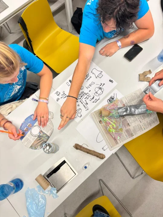 TU/e students in blue Lucid intro week shirts working together on the Salsa Shop business case, sketching ideas and building prototypes with paper, string, and recycled materials at a classroom table.