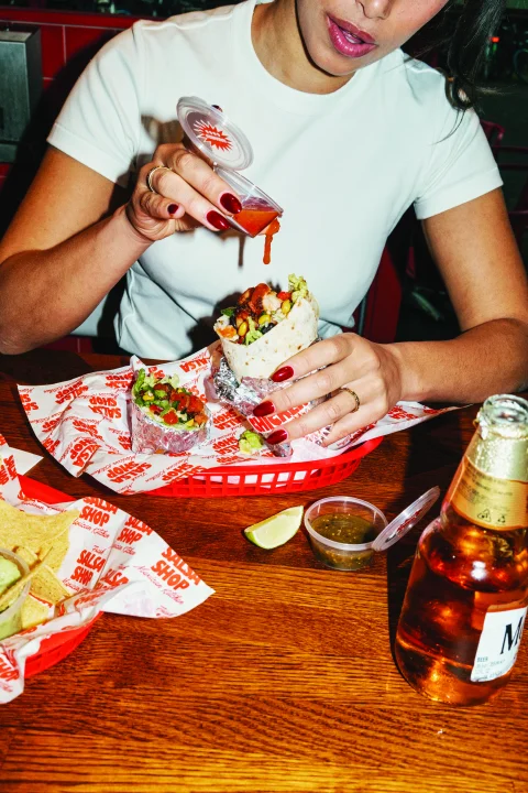 Woman adding salsa to a fresh Salsa Shop burrito at a restaurant table, with nachos, guacamole and drinks alongside.