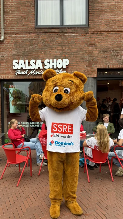 SSRE bear mascot standing outside Salsa Shop Eindhoven during introduction week, with students eating and socialising on the terrace behind.