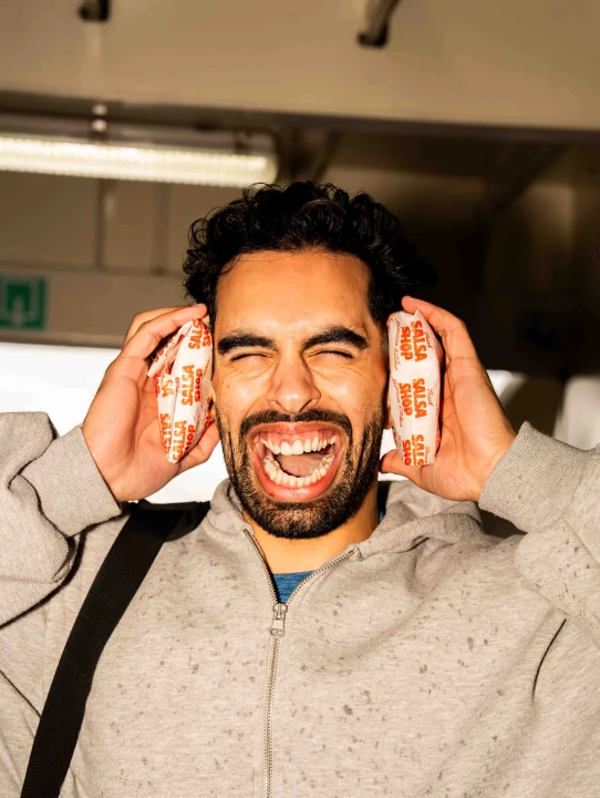 Smiling man holding two wrapped Salsa Shop burritos against his ears during a Salsa Shop campaign shoot.