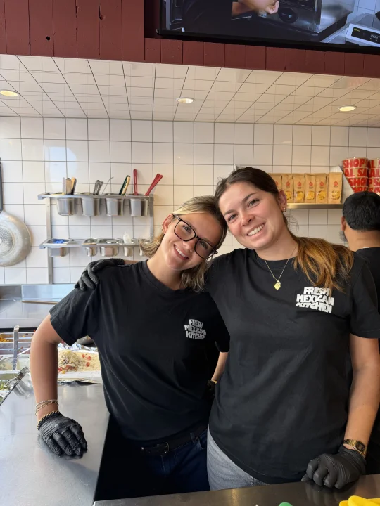 Two smiling Salsa Shop employees wearing black Fresh Mexican Kitchen T-shirts and gloves behind the counter in a restaurant.